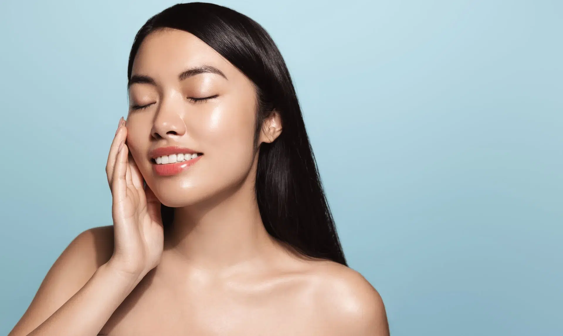 Woman with glowing skin smiling gently while touching her face against a blue backdrop.